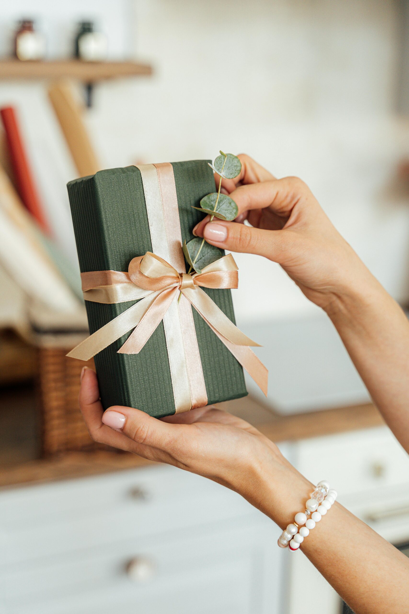 Close-up of hands holding a beautifully wrapped gift box with a ribbon and greenery accent.